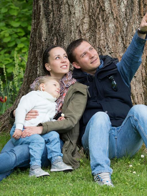 Ein junges Paar mit Kind sitzt auf einer Wiese unter einem Baum.