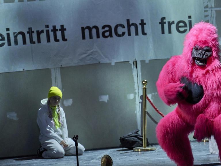 Szenenfoto: Ein Mensch verkleidet als pinkfarbener Gorilla blickt Rochtung Bühne. Im Hintergrund ein großes Plakat mit der Aufschrift: "Eintritt macht frei". Unter dem Plakat sitzt eine Jugendliche, die wohl Greta Thunberg darstellen soll.