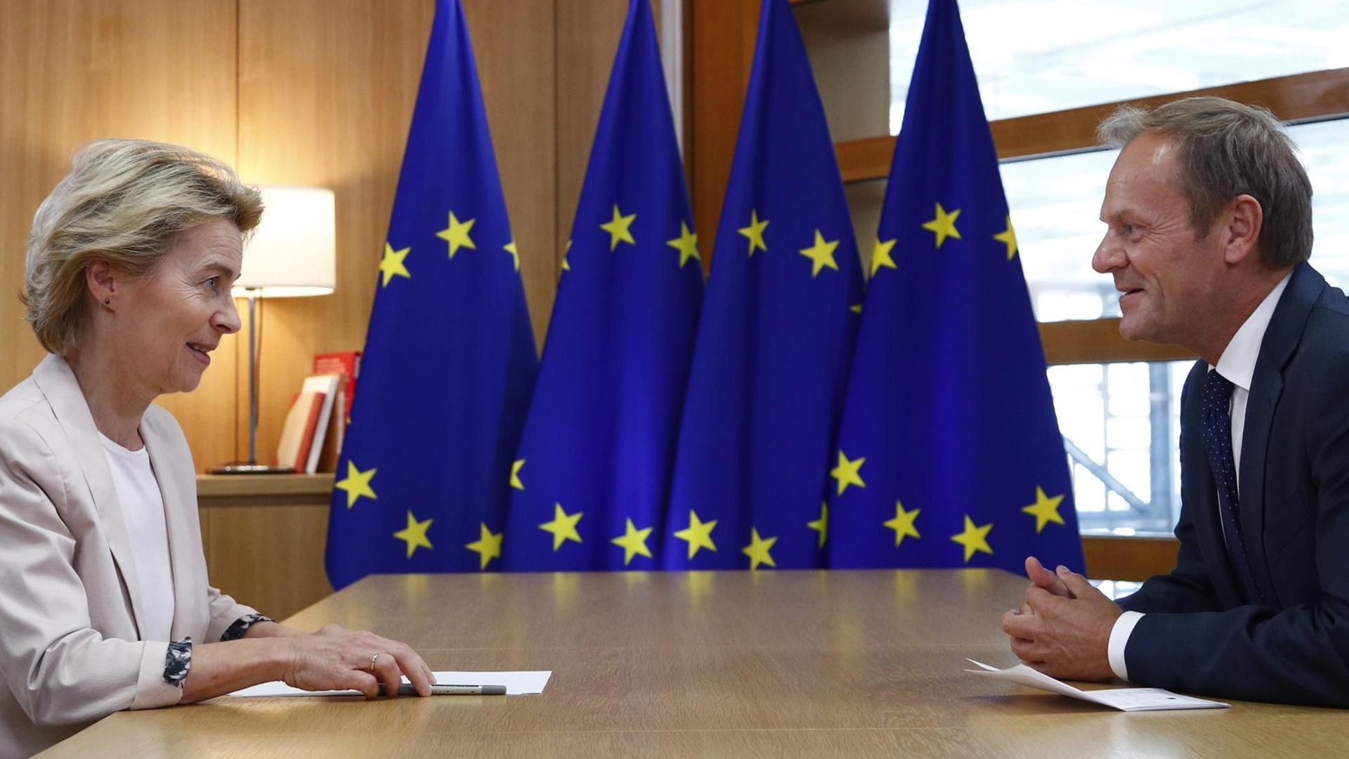 Ursula von der Leyen und Donald Tusk sitzen gemeinsam gegenüber an einem Tisch, im Hintergrund sind EU-Flaggen zu sehen.