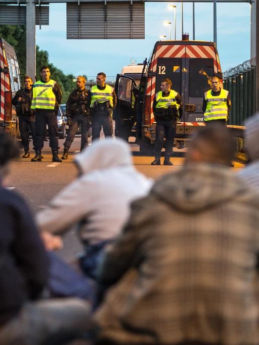 Flüchtlinge sitzen am Eurotunnel in Calais. Der Weg wird von Polizisten blockiert.