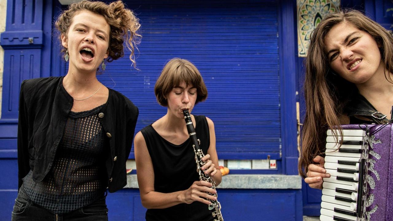 Drei Frauen stehen auf einer Straße. Eine Frau singt, die zweite Frau spielt Klarinette. Die dritte Frau hat eine Akkordeon in der Hand.