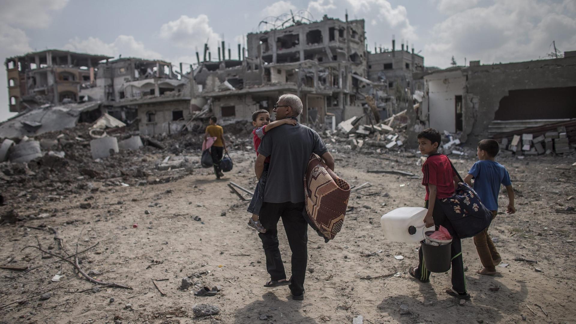 Ein Mann und seine Söhne sind auf dem Weg nach Hause, im Hintergrund sieht man zerstörte Häuser im Gazastreifen.