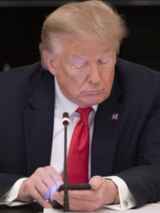 Donald Trump sitzt an einem Tisch hinter einer aufgeschlagenen Akte und einem Mikrofon und schaut auf sein Smartphone.