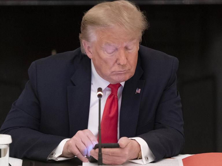 Donald Trump sitzt an einem Tisch hinter einer aufgeschlagenen Akte und einem Mikrofon und schaut auf sein Smartphone.