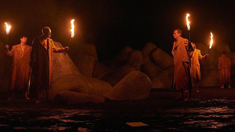 Szenenbild zur Inszenierung von "Die Hermannsschlacht" von Heinrich von Kleist Premiere am 28. November 2019. Männer tragen Fackeln in einem dunklen Bühnenbild mit großen Betonteilen.