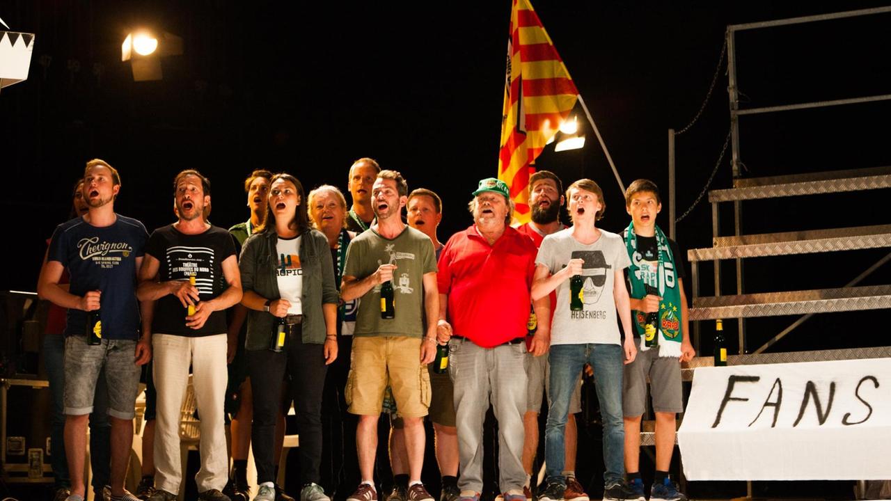 Fußballfans stehen gemeinsam mit Fahne und Bierfalsche auf der Bühne