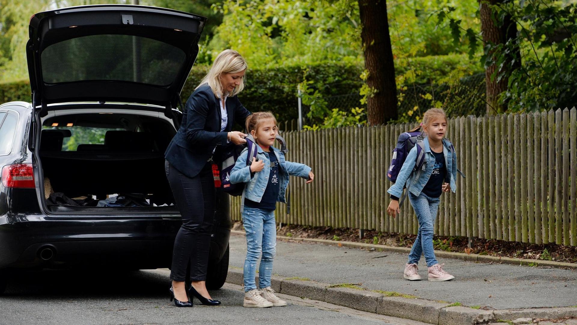 Eine Mutter in Businesskleidung hilft ihren Töchtern vor dem Auto, die Schulranzen aufzuziehen