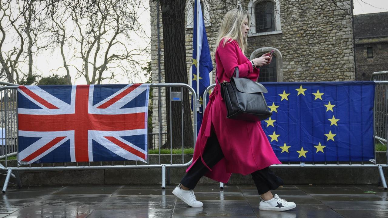 Eine Frau in einem roten Mantel geht in London an einer Absperrung vorbei, an der eine britische und eine EU-Flagge hängen.