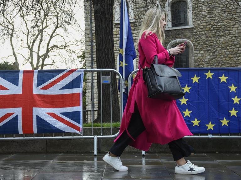 Eine Frau in einem roten Mantel geht in London an einer Absperrung vorbei, an der eine britische und eine EU-Flagge hängen.