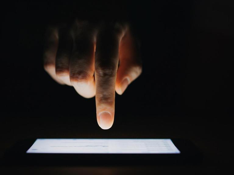 Eine Hand mit Zeigefinger tippt auf einen Bildschirm vor schwarzem Hintergrund.
