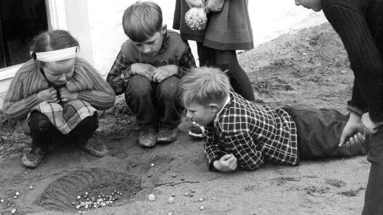 Fünf Kinder spielen im Sand mit Murmeln.