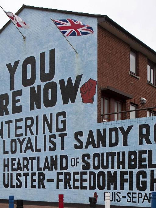 Bemalte Häuserfassade in Sandy Row, Belfast 2009: Links unten: ein maskierter und bewaffneter loyalistischer Kämpfer. Links und rechts oben: zwei geballte Fäuste, die für die Rote Hand von Ulster stehen und Symbol der UFF