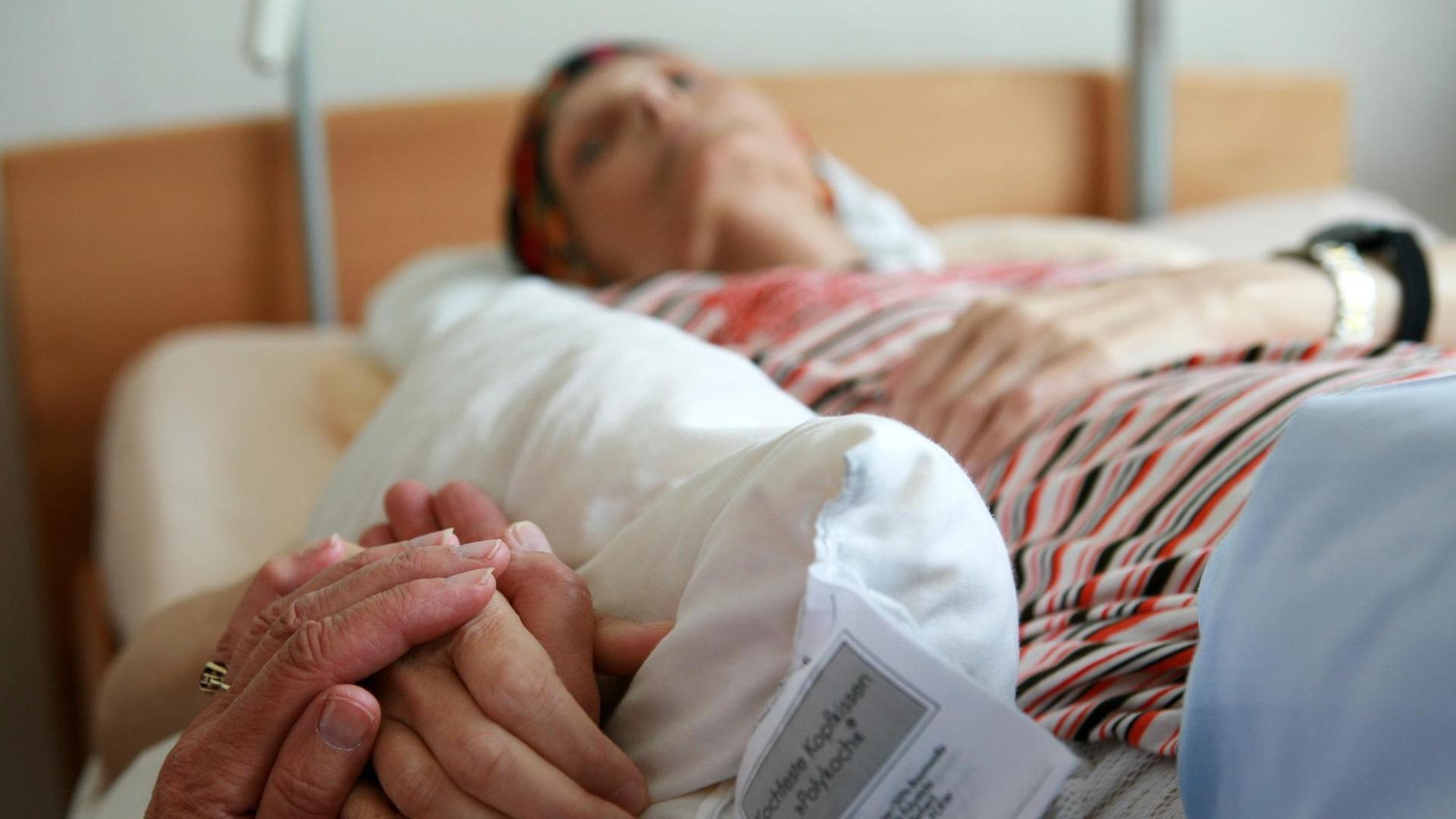 Eine ehrenamtliche Helferin hält die Hand einer sterbenden Bewohnerin, die in ihrem Bett im Christophorus Hospiz in München liegt