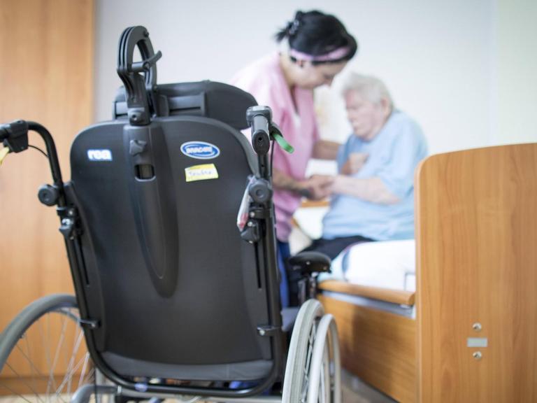 Eine Altenpflegerin hilft einer Bewohnerin in einem Altenpflegeheim aus dem Bett. Im Vordergrund steht ein Rollstuhl.