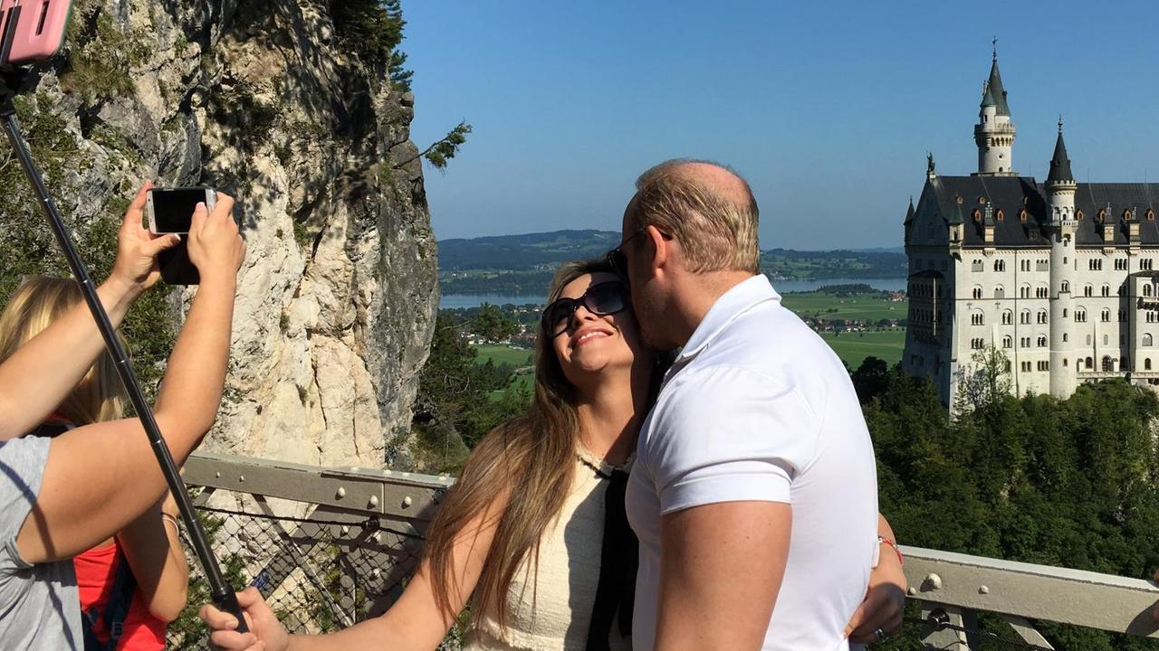 Ein Paar macht ein Selfie von sich. Im Hintergrund das Schloss Neuschwanstein. 