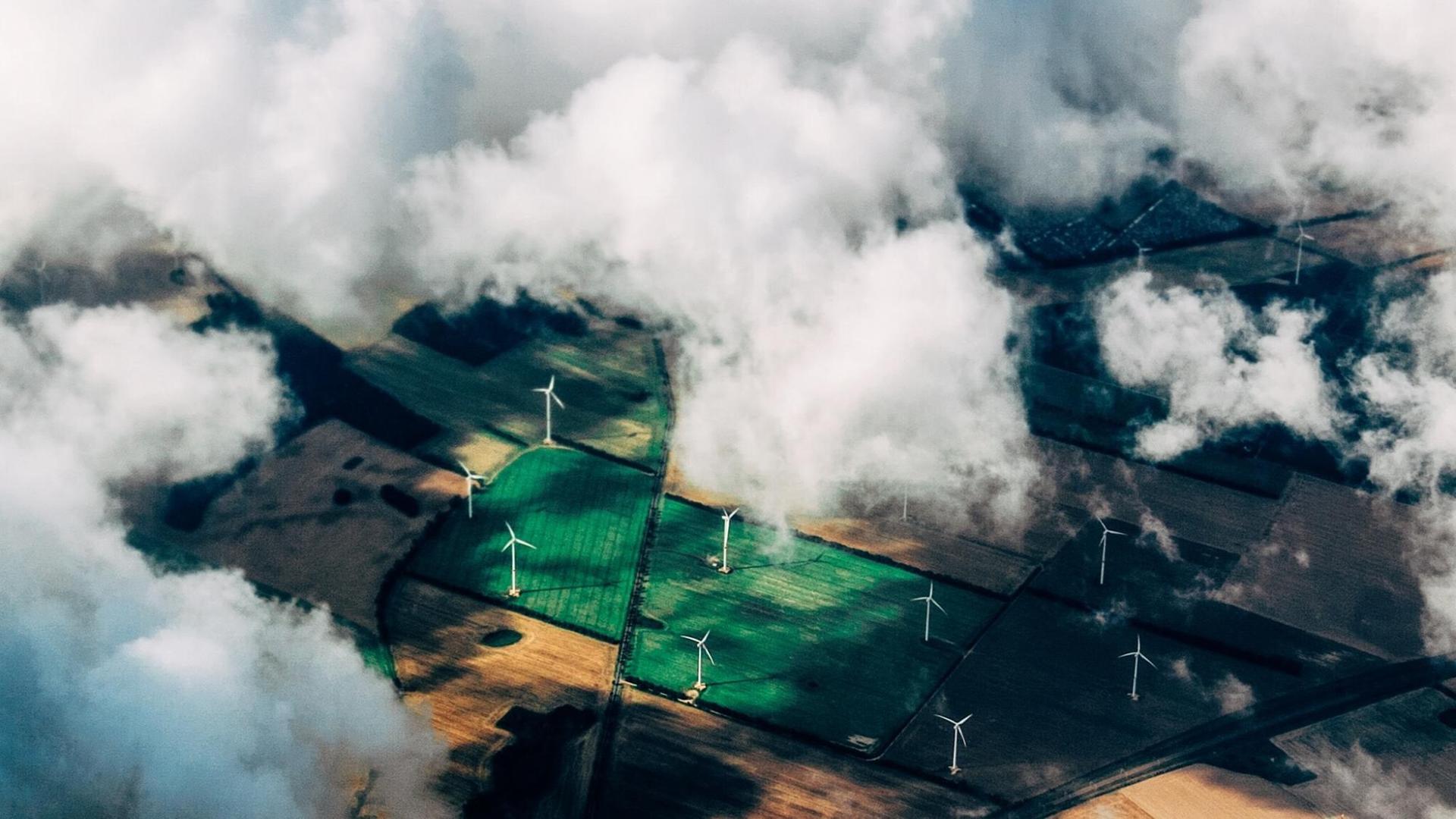 Eine Luftaufnahme zeigt Felder in verschiedenen Grüntönen mit Windkraftanlagen zwischen einem dramatischem Himmel.