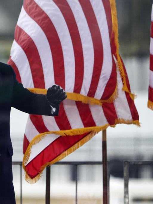 Trump steht hinter Glasscheiben vor der amerikanischen Flagge und ballt seine Hände zu Fäusten.