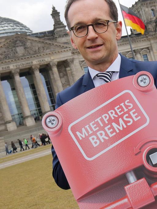 Heiko Maas steht auf der Wiese vor dem Reichstag und hält ein Schild in Händen, das eine Notbremse darstellt und auf dem "Mietpreisbremse" steht.