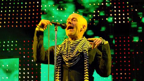Konzert der Band R.E.M in der Berliner Waldbühne im Jahr 2008. Sänger Michael Stipe steht auf der Bühne am Mikro vor einer großen Leinwand und singt, er trägt einen auffälligen, geringelten Schal