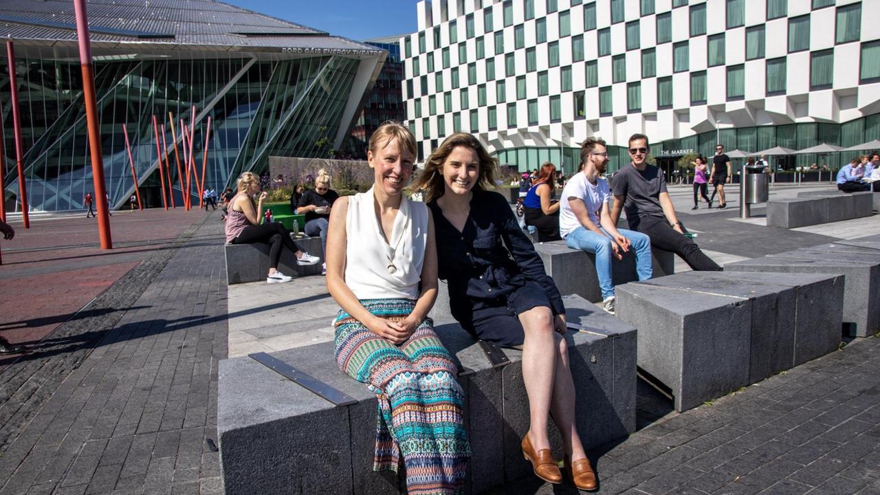 Zwei Frauen sitzen bei Sonnenschein auf einem Steinblock in der Mittagspause vor Bürogebäuden aus Glas, Stahl und Beton.