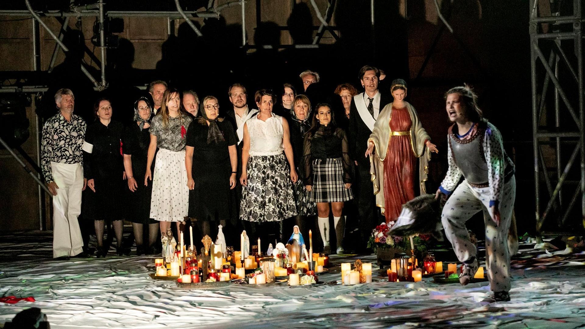 Szenenfoto aus "Überwältigung" von Thomas Melle bei den Nibelungen Festspielen Worms, in dem der Chor des Nibelungen im Hintergrund zu sehen ist, während vorne rechts Ortlieb gespielt von Lisa Hrdina schreit und mit dem Fuss aufstampft.