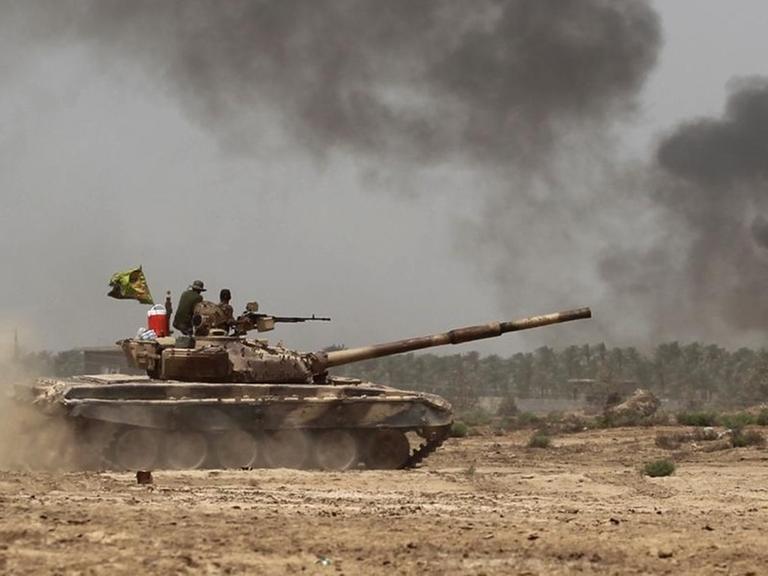 Ein Panzer fährt durch sandiges Gelände, im Hintergrund schwarzer Rauch.