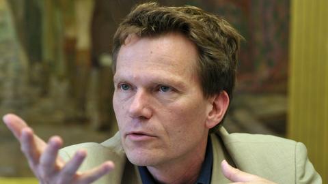 Ein mittelalter Mann mit dunkelblondem kurzen Haar und blaugrauen Augen gestikuliert beim Reden mit den Händen. Er trägt ein beiges Jackett, im Hintergrund ist unscharf ein Gemälde zu erkennen.