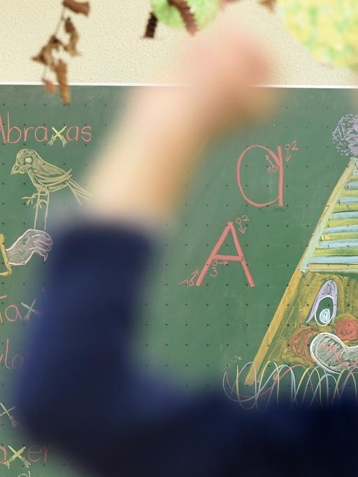 Ein Junge wirft in einem Klassenzimmer mit einem Gegenstand. Im Hintergrund ist ein Tafelbild mit einer Hexe.