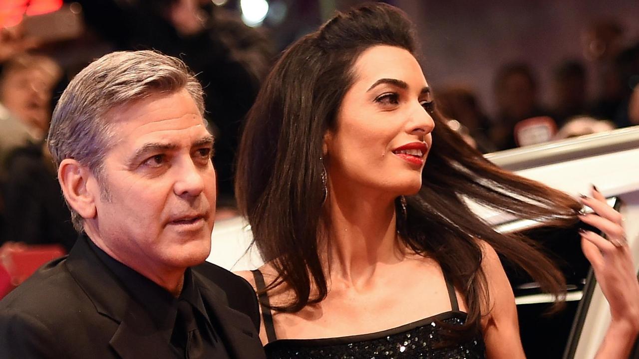 Clooney und Amal schauen engumschlungen in die Kameras, Amal zieht an ihren Haaren. Er trägt einen schwarzen Anzug, sie ein schwarzes schulterfreies Kleid.