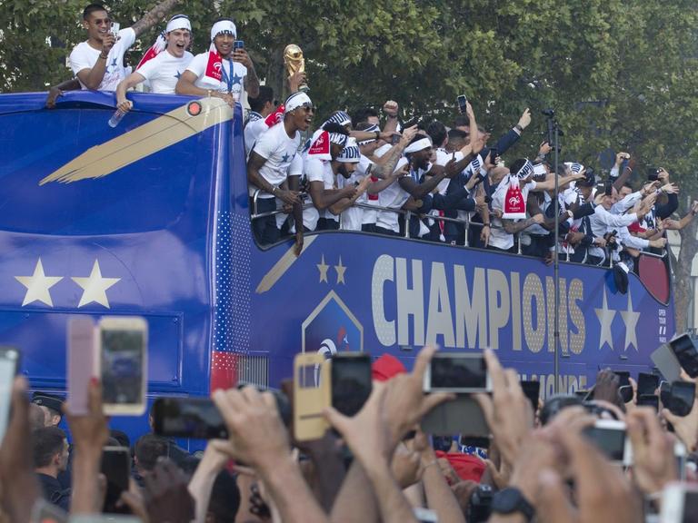 Die Spieler stehen auf einem offenen Doppeldecker-Bus mit der Aufschrift "Champions". Sie tragen weiße T-Shirts und winken den jubelnden Zuschauern zu.