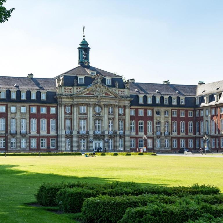 Rotes Schloss, im Vordergrund eine grüne Wiese, auf der eine kleine Gruppe Menschen sitzt.