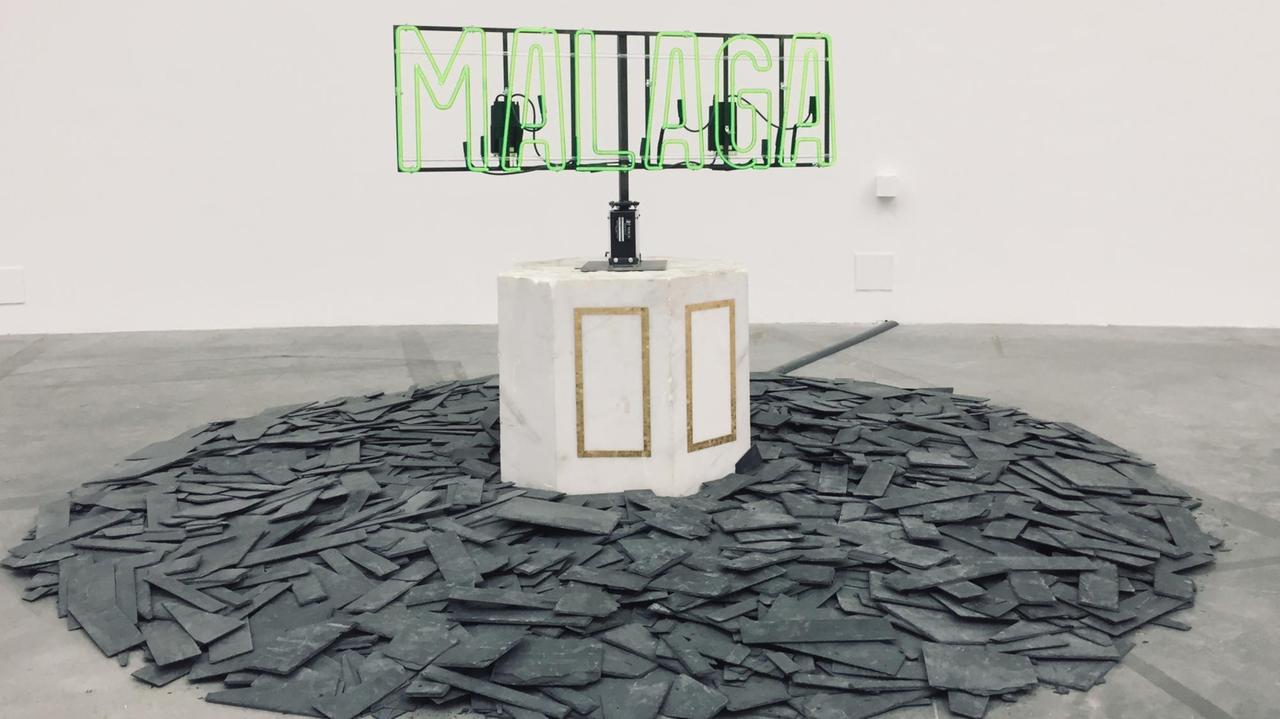 Ein Werk aus der Ausstellung "Amalgam" des US-Amerikaners Theaster Gates im Palais de Tokyo Paris. Zerschlagene Schieferplatten auf einem Haufen, in der Mitte ein Marmorblock mit einem Metallgestänge, auf dem eine neongrüne Leuchtschrift steht: zu lesen ist "MALAGA".