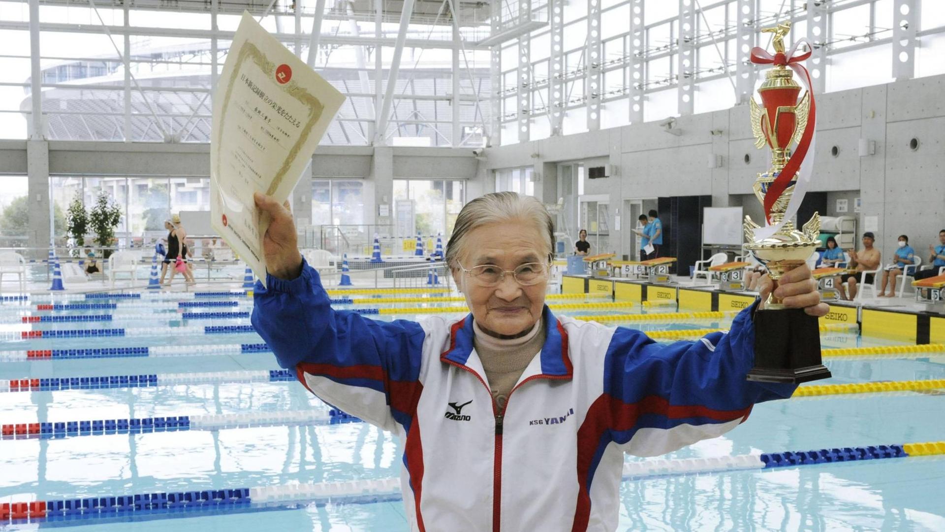 Eine Hundertjährige Japanerin feiert ihren Titelgewinn bei einem Schwimm-Wettbewerb. Sie hält eine Urkunde und einen Pokal in die Höhe, im Hintergrund ist das Wettkampf-Schwimmbecken zu sehen. Eine Hundertjährige Japanerin feiert ihren Titelgewinn bei einem Schwimm-Wettbewerb. Sie hält eine Urkunde und einen Pokal in die Höhe, im Hintergrund ist das Wettkampf-Schwimmbecken zu sehen.