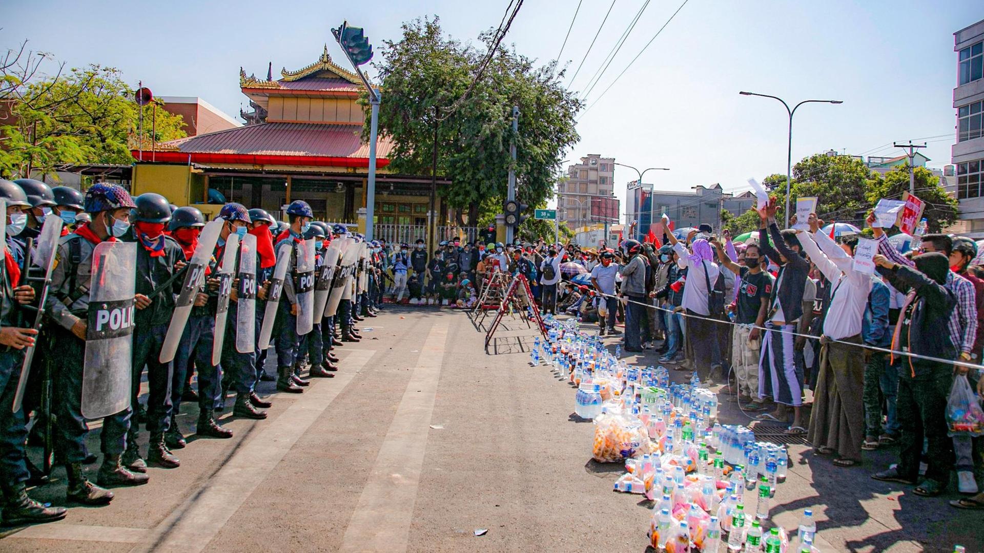 Polizisten und Demonstranten stehen sich an einer Straße in der Stadt Mandalay in Myanmar gegenüber. Die Protestierenden habe Wasserflaschen hinter die Absperrung gestellt und halten Schilder in die Höhe.