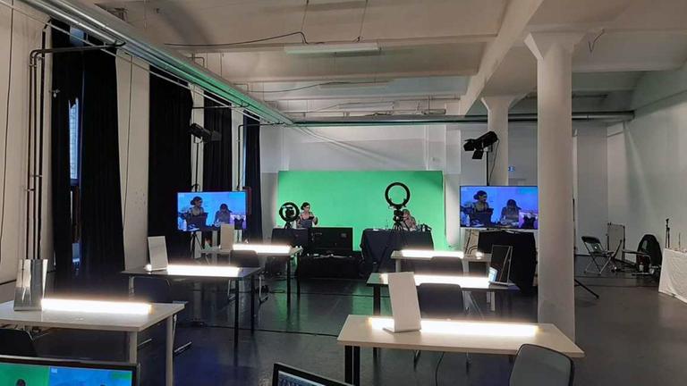Aufnahme vom digitalen Facefilter-Workshop am Theater Hebbel am Ufer in Berlin. In einem Arbeitsraum mit beleuchtet Tischen, einem Green-Screen und zwei Personen vor der Kamera zu sehen.
