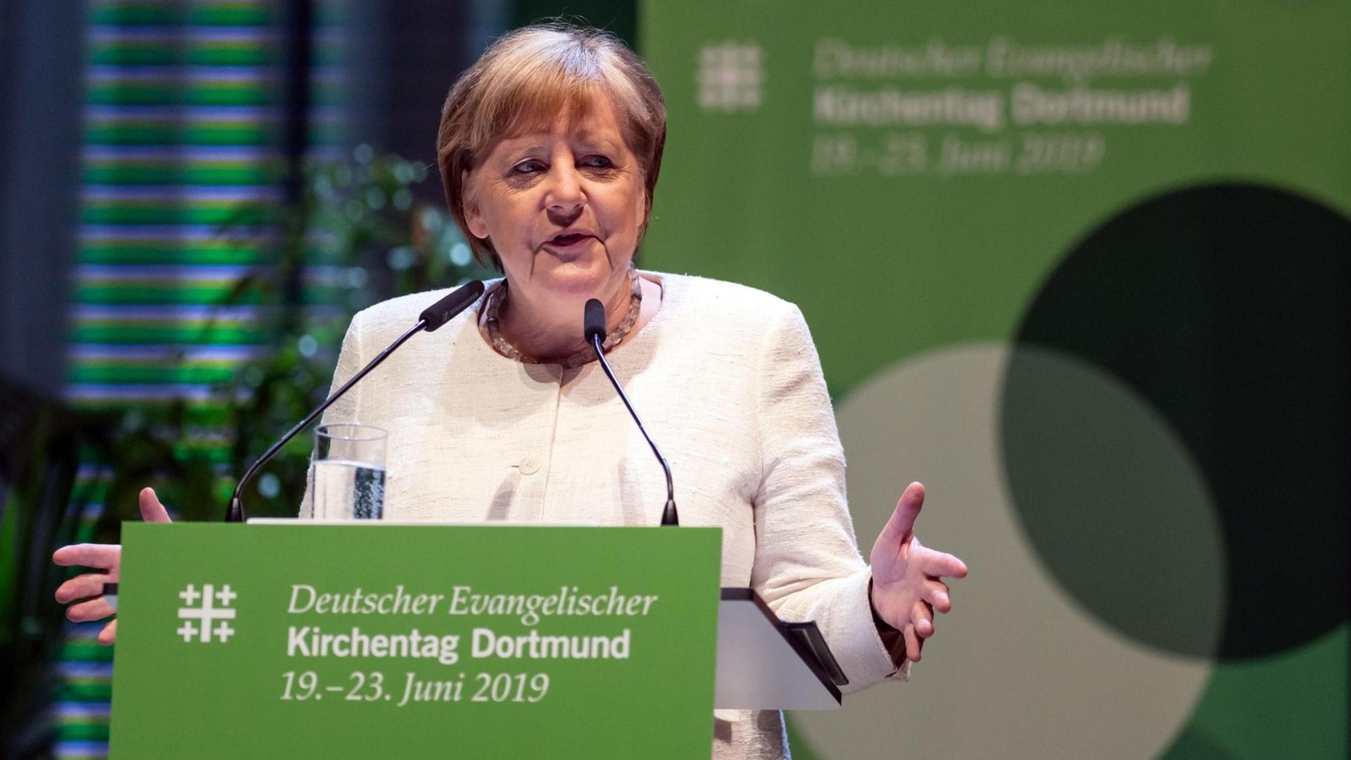 Merkel steht an einem Redner-Pult mit einem grünen Schild von dem Kirchentag. Sie spricht und breitet dabei beide Hände aus.