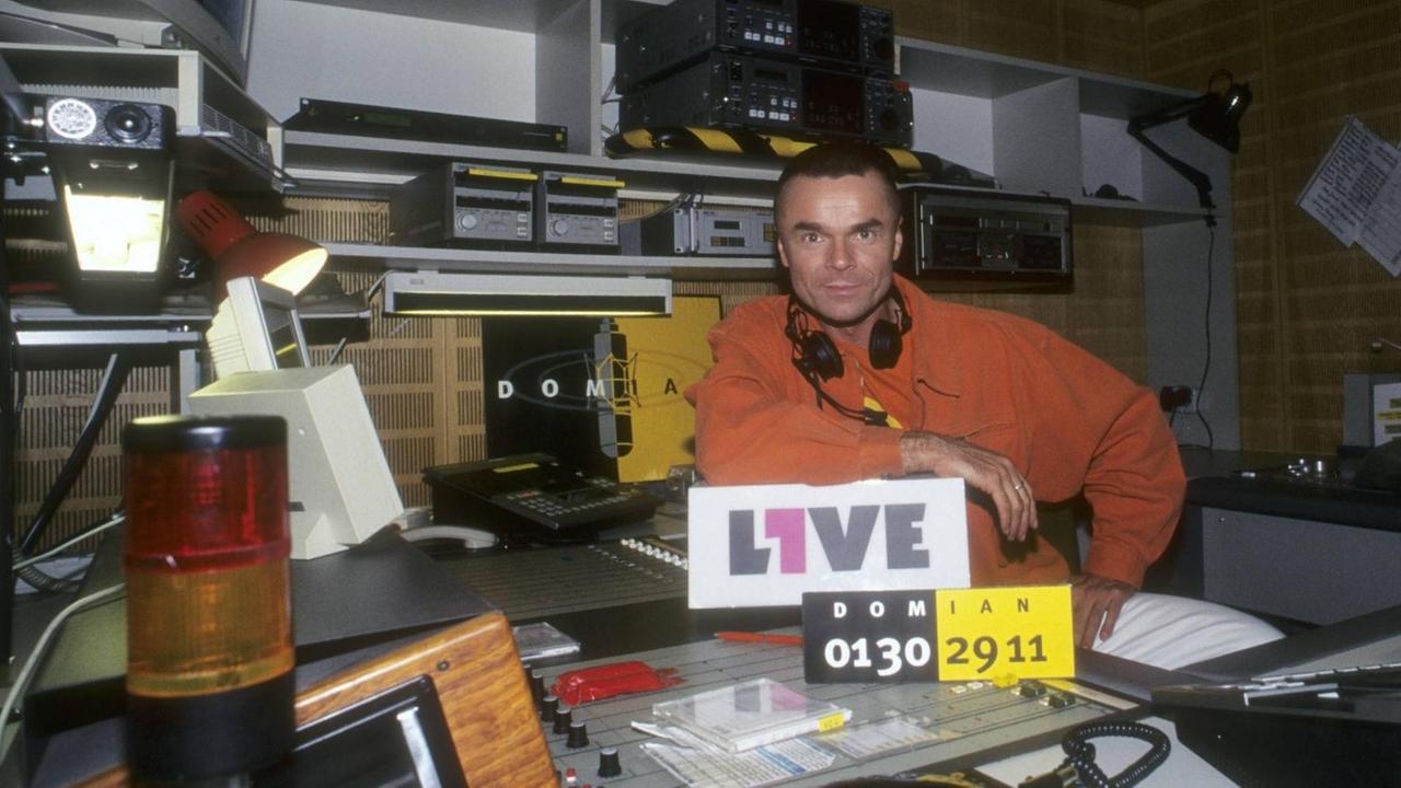 Jürgen Domian sitzt 1996 im 1Live-Studio, von dem erseine Sendung "Domian" moderierte  - im Vordergrund ein Schild mit der Telefonnummer für die Hörerinnen und Hörer