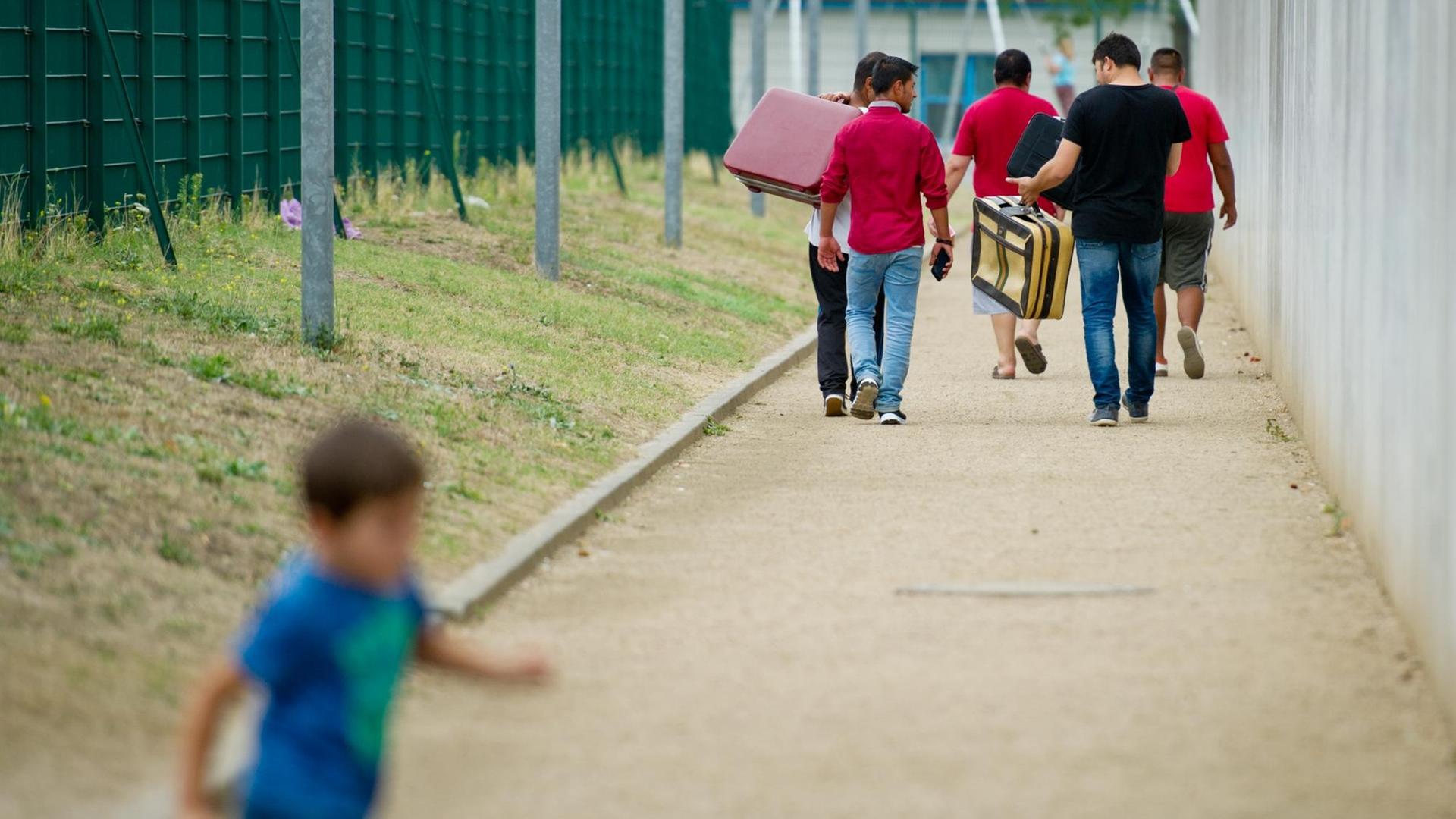 Flüchtlinge laufen am 27.08.2015 mit Koffern bepackt auf einem Weg einer Erstaufnahmeeinrichtung für Flüchtlinge in Ingelheim (Rheinland-Pfalz) entlang, während ein Kind im Vordergrund vorbeiläuft. 