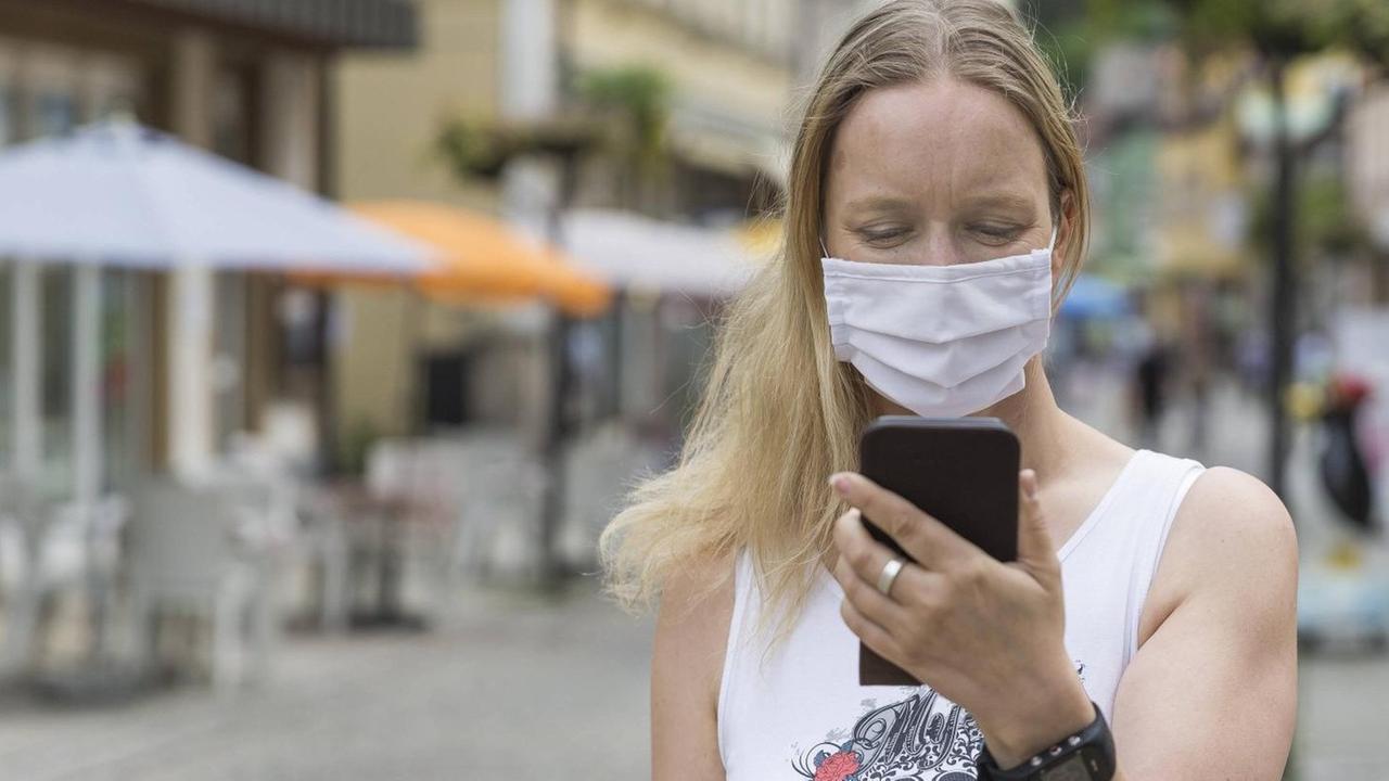 Eine junge Frau mit Mund-Nasen-Behelfs-Schutz schaut in einer Fußgängerzone auf ihr Handy.