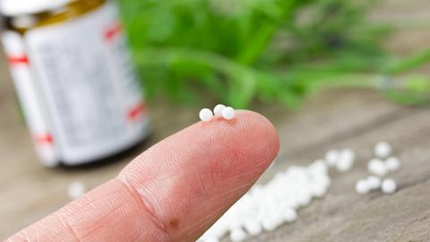 Auf einer Fingerspitze liegen drei weiße Kügelchen, im Hintergrund liegen weitere Kügelchen auf einer Holzplatte, in der linken Ecke ist eine kleine Arzneiflasche zu sehen