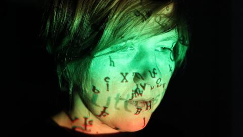 Portrait einer Frau mit schwarzen Haaren. Sie ist grün und orange angestrahlt. Auf dem Gesicht spiegeln sich Buchstaben.