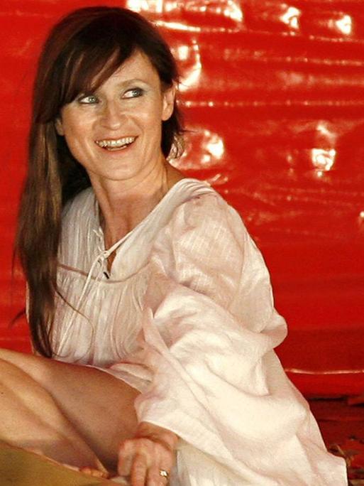 Eine braunhaarige Schauspielerin sitzt lächelnd vor einer grellroter Wand.