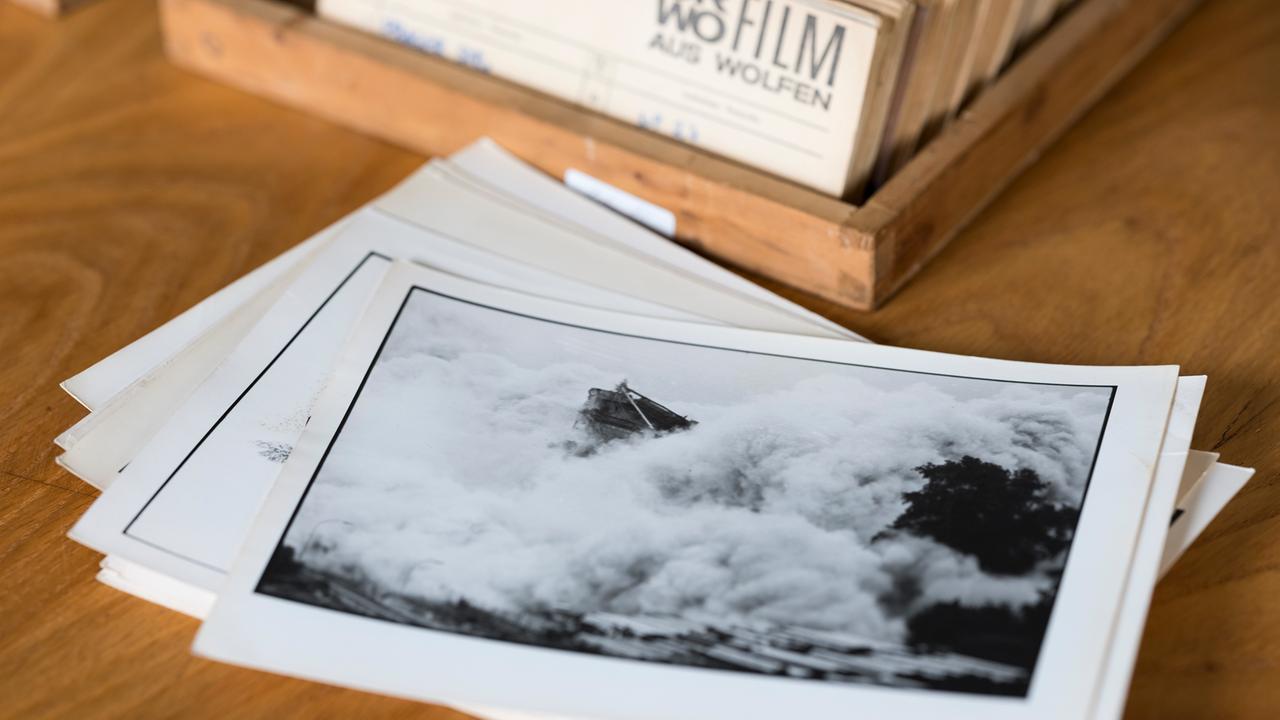 Auf einem Holztisch liegen ein Stapel Schwarz-Weiß-Fotos, im Hintergrund ein Zettelkasten voller Negative oder Fotos. Das oberste Foto zeigt einen Fels in der Brandung.