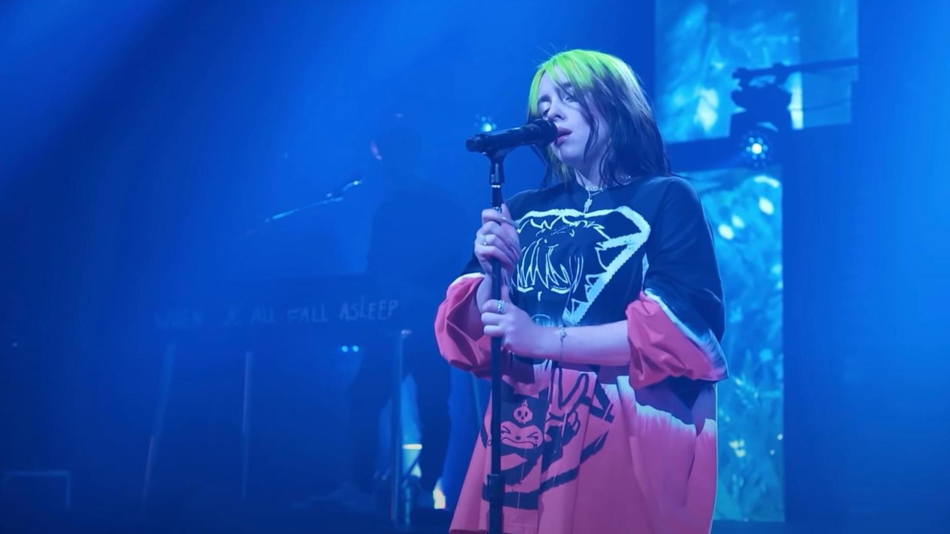 Billie Eilish steht singend auf einer Bühne und umklammert mit den Händen den Sänder des Mikrofons. Sie trägt ein sehr weites, sackartiges und gemustertes Oberteil. Ihre Haare sind schwarz und neon-grün gefärbt.