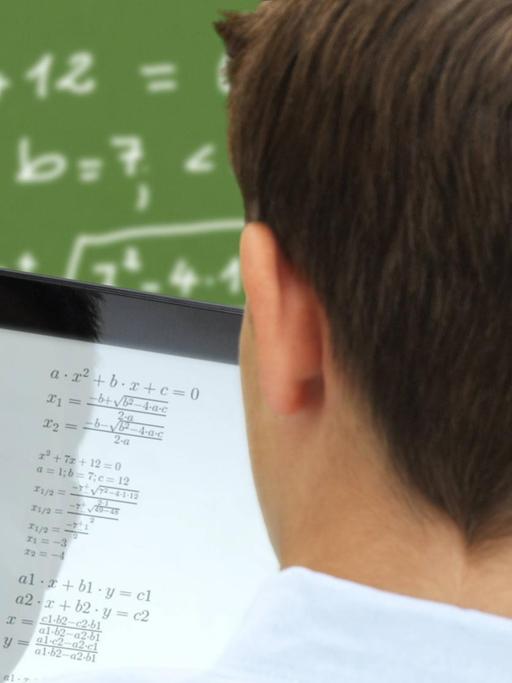 Ein Schüler mit einem Tabletcomputer steht vor einer Schultafel mit mathematischer Gleichung