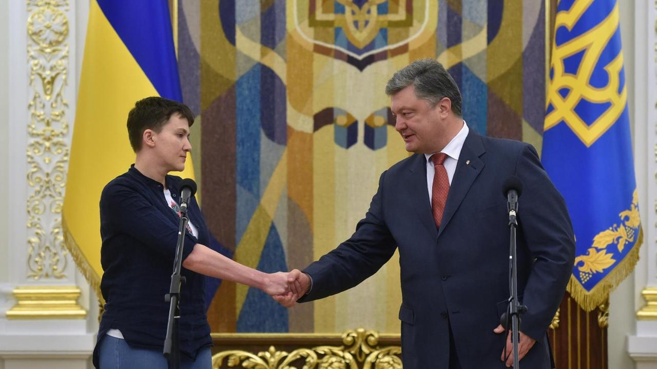 Nadja Sawtschenko und Petro Poroschenko stehen vor Mikrofonen und geben sich die Hand. Im Hintergrund ukrainische Flaggen.