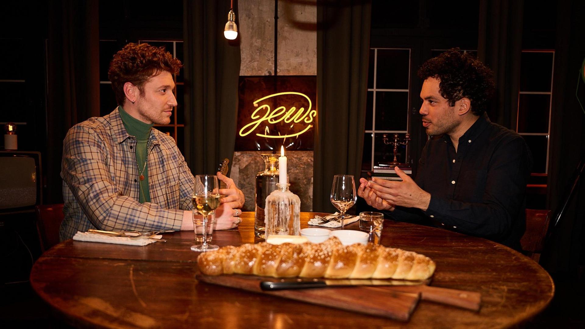 Zwei junge Männer sitzen an einem runden, gedeckten Esstisch aus Holz und unterhalten sich. Hinter ihnen befindet sich als gelbe Leuchtschrift das Wort "Jews".