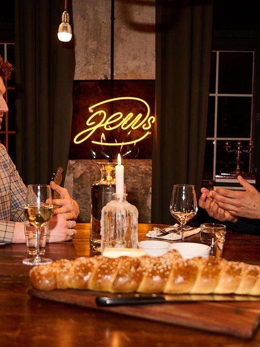 Zwei junge Männer sitzen an einem runden, gedeckten Esstisch aus Holz und unterhalten sich. Hinter ihnen befindet sich als gelbe Leuchtschrift das Wort "Jews". Zwei junge Männer sitzen an einem runden, gedeckten Esstisch aus Holz und unterhalten sich. Hinter ihnen befindet sich als gelbe Leuchtschrift das Wort "Jews".