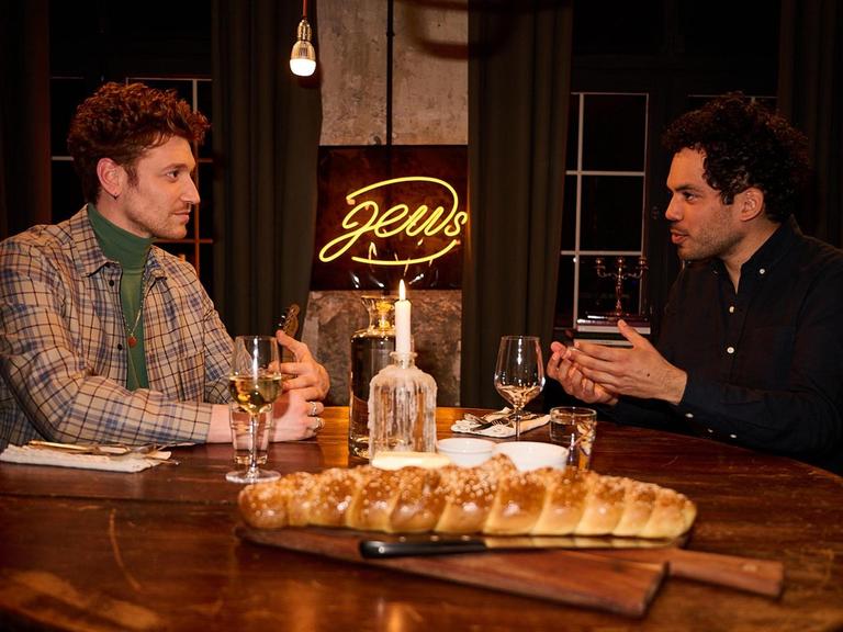 Zwei junge Männer sitzen an einem runden, gedeckten Esstisch aus Holz und unterhalten sich. Hinter ihnen befindet sich als gelbe Leuchtschrift das Wort "Jews". Zwei junge Männer sitzen an einem runden, gedeckten Esstisch aus Holz und unterhalten sich. Hinter ihnen befindet sich als gelbe Leuchtschrift das Wort "Jews".
