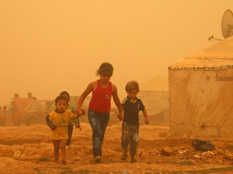 Zu sehen sind drei Kinder in einem Flüchtlingslager bei Baalbek im Libanon, die Luft ist wegen eines Sandsturms gelb.
